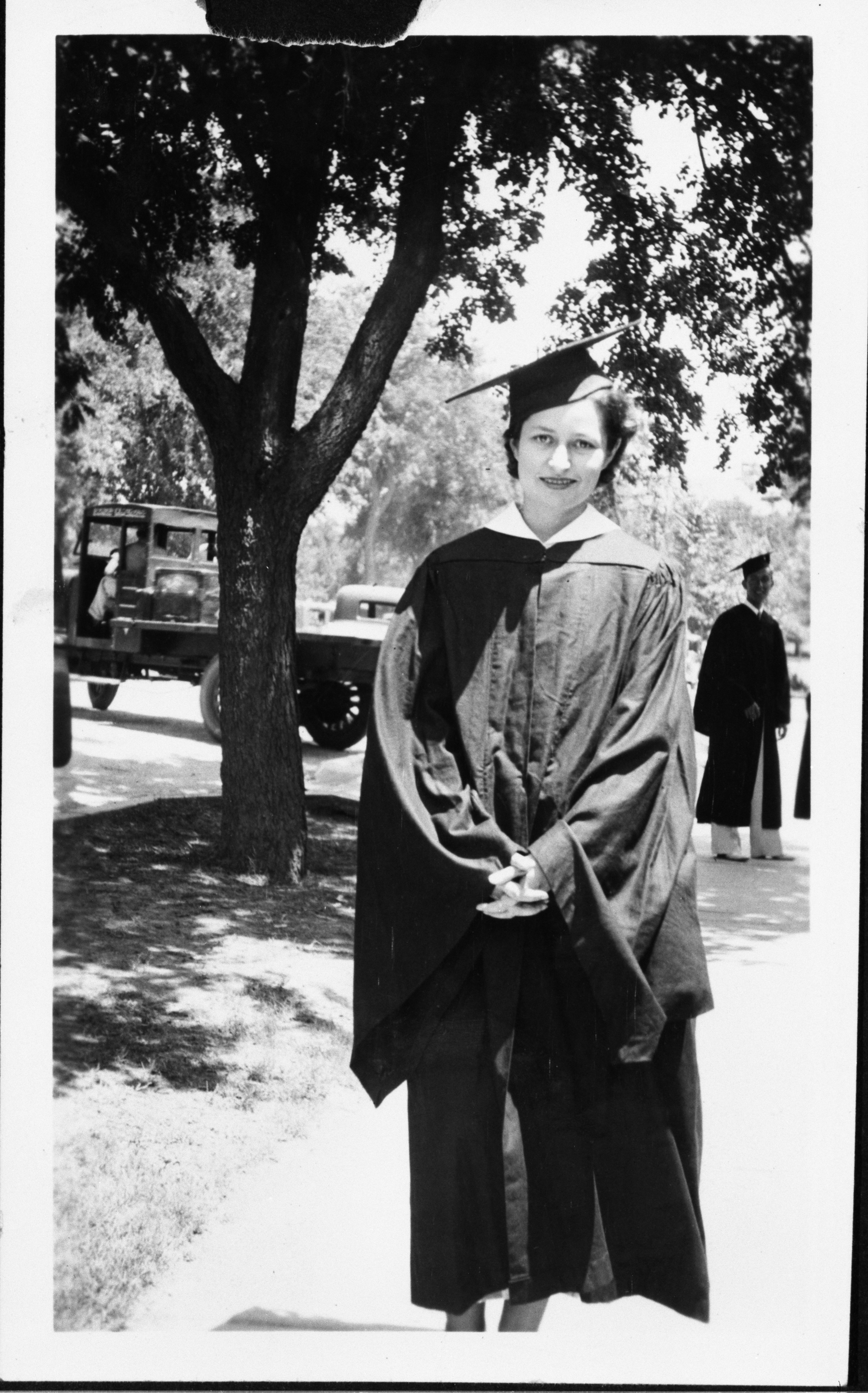 Claudia "Lady Bird" Taylor stands in cap and gown on sidewalk with a tree and truck in background